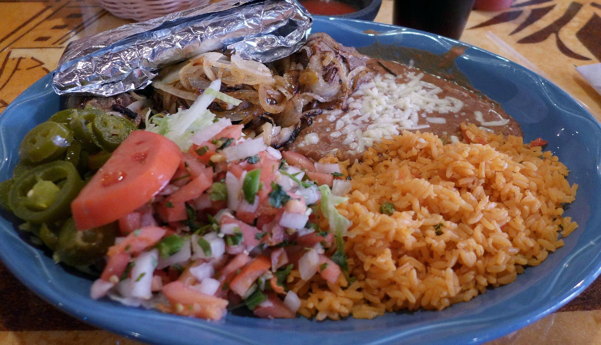 A carne asada plate — grilled steak topped with sautéed onions, with rice, refried beans, pico de gallo, jalapeños and tortillas in foil